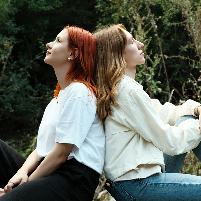 En bild på två unga kvinnor rygg mot rygg som tittar upp mot himmelen
